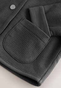 Gray textured fabric featuring a pocket and fitted cuff. The design includes a smooth finish at the edges and a button detail.