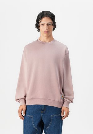 Lyserød sweatshirt i blødt stof med rund halsudskæring og batwingærmer. Den har et logo-patch i nederste venstre side, sammen med blå jeans.