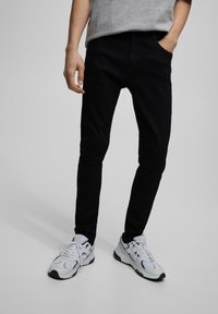 Jeans negros de corte ajustado con una textura suave, combinados con zapatillas deportivas blancas que presentan detalles de malla y goma. Fondo en escala de grises.