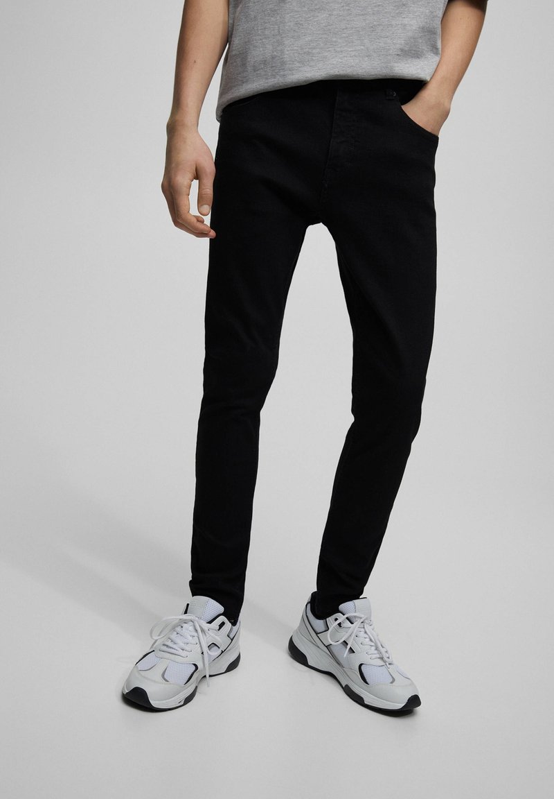 Jeans negros de corte ajustado con una textura suave, combinados con zapatillas deportivas blancas que presentan detalles de malla y goma. Fondo en escala de grises.