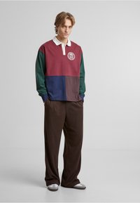 Langærmet polo i marmor, grøn, navy og mørkebrun farver med en hvid krave og cirkulært logo. Løst siddende brune bukser, grå slippers.