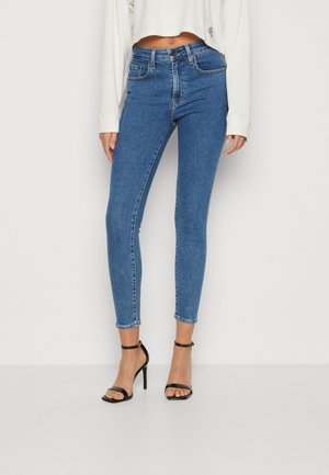 Femme portant un jean skinny bleu taille haute, un sweat-shirt court blanc et des sandales à talons hauts avec bride à la cheville noires, debout contre un mur blanc.