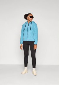 Giacca in pile azzurro chiaro con cappuccio, due tasche anteriori e dettagli con cerniera nera, abbinata a leggings neri e sneakers beige.