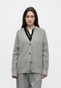 Šedý pletený kardigan s texturovaným vzorem, zapínáním na knoflíky vpředu a výstřihem do V. Nošený přes černou košili s bílými pruhy.