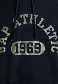 Sudadera negra con texto desgastado en verde que dice "GAP ATHLETIC 1969", con capucha ajustable y tejido texturizado.