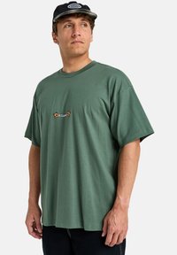 Green cotton t-shirt with a relaxed fit, short sleeves, and a small embroidered "Billabong" logo with flower accents on the chest.