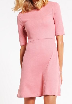 Woman wearing a fitted, ribbed, dusty pink dress with elbow-length sleeves and a slightly flared skirt against a white background.