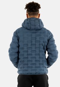 Giacca a vento imbottita con cappuccio di un blu intenso, caratterizzata da una texture trapuntata con sezioni rettangolari e polsini elasticizzati, abbinata a pantaloni neri.