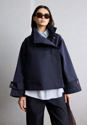 Femme portant une veste bleu marine oversize avec de larges poignets et des lunettes de soleil écaille, tenant un sac texturé marron foncé.