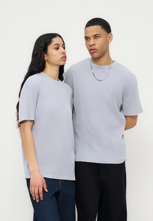 Δύο άτομα φοράνε ανοιχτογκρι κοντομάνικα πουκάμισα, στέκονται δίπλα δίπλα μπροστά σε ένα απλό λευκό φόντο.