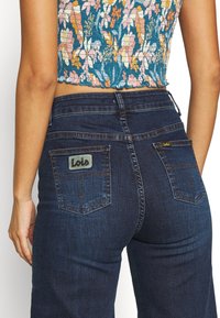 Tmavě modré denimové džíny s vysokým pasem, které mají dvě zadní kapsy a stříbrnou a černou etiketu "Lois" přišitou na pravé kapse.