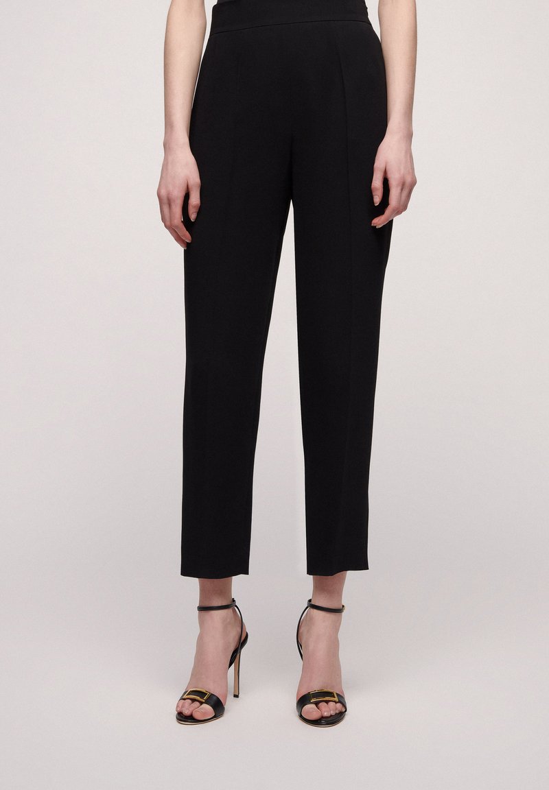 Pantalons noirs ajustés avec une taille haute, des jambes droites et de légères plis à l'avant. Associés à des sandales à talons hauts noires à lanières.