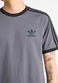 Gray Adidas t-shirt with black collar and three striped sleeves, featuring an embroidered logo on the chest and smooth fabric texture.