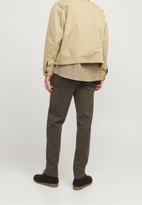 Homme portant une veste beige, une chemise à carreaux, un pantalon marron foncé et des chaussures noires, debout dos au spectateur sur un fond uni.