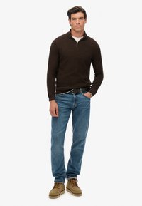 Bruine gebreide trui met kwart-rits, blauwe vervaagde jeans en beige suède laarzen met veters. Model staat zelfverzekerd met de handen casual geplaatst.