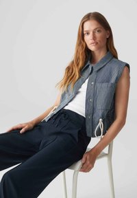Gilet a righe senza maniche in blu navy e bianco con colletto, tasca frontale e coulisse; abbinato a pantaloni blu navy. Modella seduta su una sedia.