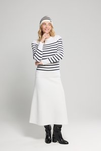 Vrouw in een witte rok, zwarte laarzen, wit langemouwen shirt met zwarte horizontale strepen en bijpassende gestreepte beanie, staande tegen een effen achtergrond.