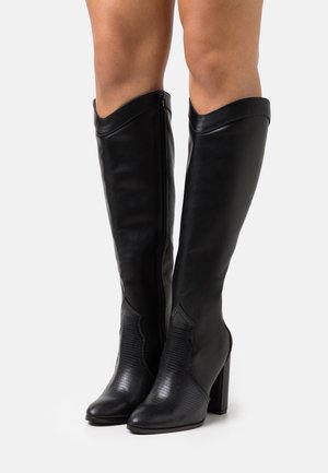 Bottes en cuir noires montantes jusqu'aux genoux, avec bouts pointus, panneaux avant texturés, fermetures éclair latérales et talons carrés, portées sur des jambes nues.