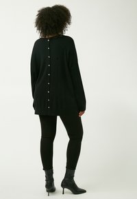 Schwarzer, übergroßer Pullover mit asymmetrischem Saum, der auf der Rückseite eine Reihe dekorativer Knöpfe aufweist, sowie langen Ärmeln, kombiniert mit schwarzen Leggings.