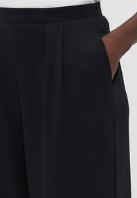 Schwarze Hosen mit glatter Textur, hohem Bund, vorderen Falten und seitlichen Taschen. Der Stoff wirkt leicht und hängt fließend.