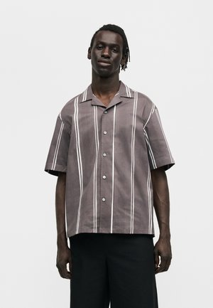 Άνδρας που φοράει φαρδιά γκρι κοντομάνικη μπλούζα με κάθετες λευκές ρίγες και μαύρο παντελόνι, στέκεται μπροστά σε απλό ανοιχτό φόντο.