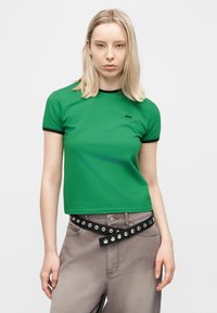 Νεαρή γυναίκα με ίσια ξανθά μαλλιά, που φοράει πράσινο μπλουζάκι με μαύρη επένδυση και γκρι παντελόνι με μαύρη ζώνη με τρουκς.