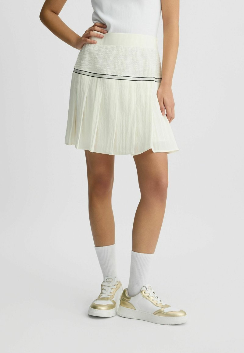 Femme portant une jupe plissée blanche avec une ceinture texturée, des chaussettes blanches et des baskets blanches à accents dorés, debout avec une main sur la hanche.