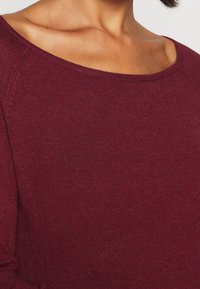 Maglione borgogna con un morbido tessuto a maglia testurizzato, scollo a barca ampio e maniche raglan, presenta un colore uniforme senza motivi.