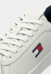 Witte synthetische sneaker met platte veters, een gestructureerde tong en een klein rood, wit en blauw logo-label met de tekst "TOMMY JEANS" aan de zijkant.
