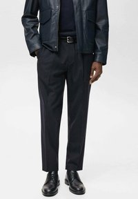 Lederjacke mit Fronttaschen, dunkelblaue Hose und schwarze Oxfordschuhe. Schlichter, taillierter Schnitt mit glatter Textur und klaren Linien.
