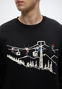 Μαύρο φούτερ με λευκή κεντητή σχεδίαση σκηνών σκι, με μπλε, κόκκινες και πορτοκαλί λεπτομέρειες, σε φόντο βουνού.