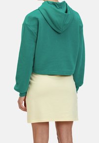 Una persona che indossa un hoodie verde corto e una gonna gialla, vista di spalle, si trova davanti a uno sfondo neutro.