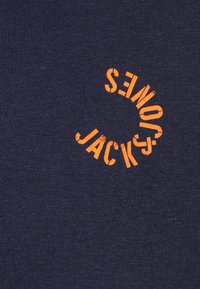 Camiseta de algodón azul marino con texto curvo en naranja que dice "JACKSON'S." La tela tiene una textura suave con un diseño minimalista.