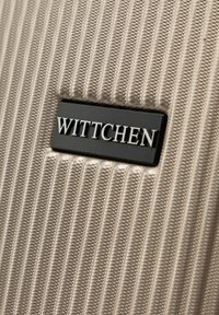 Zbliżenie na beżową teksturowaną powierzchnię walizki z czarną prostokątną metką z nazwą marki "WITTCHEN" w srebrnych literach.