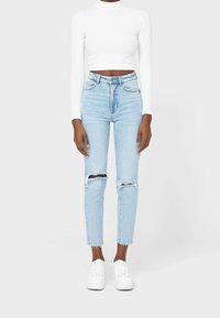 Light blue high-waisted jeans with distressed knees, paired with a fitted white long-sleeve top and white sneakers. Classic denim texture.