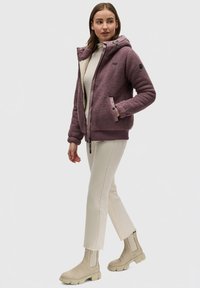 Veste à capuche en polaire de couleur violet discret, avec un intérieur clair contrastant et une fermeture éclair devant. Associée à un pantalon crème et des bottines beiges.