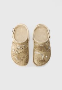 Beige Clogs mit strukturiertem Canvas-Obermaterial, das gestickte Muster und dekorative Silbernieten aufweist. Ergonomisches Fußbett und verstellbarer Riemen.