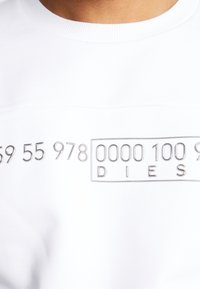 Bílá mikina s hladkou texturou, na které jsou na přední straně kovově stříbrné vyražené číslice a slovo "DIESEL". Kulatý výstřih.