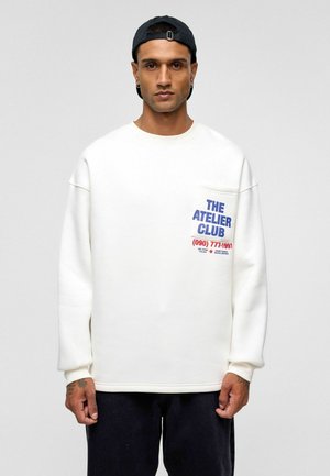 Witte sweatshirt met een ontspannen pasvorm, met een borstzak en meerkleurige tekst. Model draagt donkere broek en een zwarte pet.