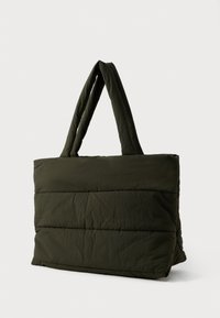 Stor grønn polstret toteveske med brede håndtak, som står oppreist på en hvit bakgrunn.