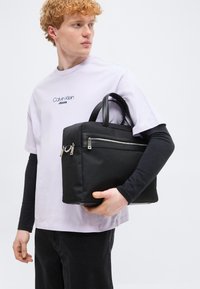 Νεαρός άνδρας με σγουρά κόκκινα μαλλιά που φοράει λευκό πουκάμισο Calvin Klein με μαύρα μανίκια, κρατώντας μια μαύρη τσάντα χειρός με φερμουάρ.