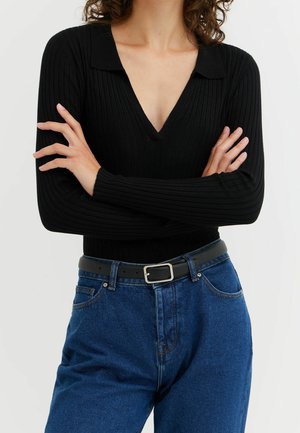 Personne aux cheveux bouclés portant un haut noir côtelé à manches longues et un jean bleu avec une ceinture noire, les bras croisés sur la poitrine.