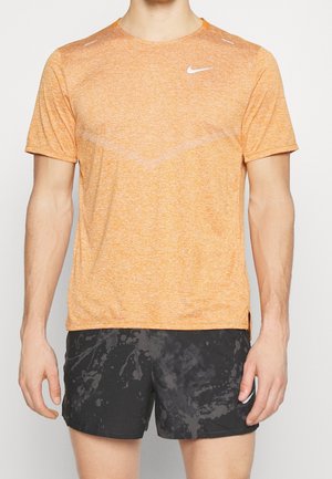 Camiseta atlética naranja de Nike de manga corta con tejido texturizado, detalles reflectantes en los hombros, combinada con pantalones cortos negros con un patrón salpicado.