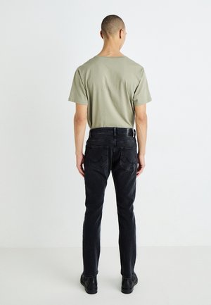 Ein Mann steht mit dem Rücken zur Kamera, trägt ein hellgrünes kurzärmeliges Shirt, schwarze Jeans mit Gesäßtaschen und schwarze Schuhe vor einem weißen, einfarbigen Hintergrund.