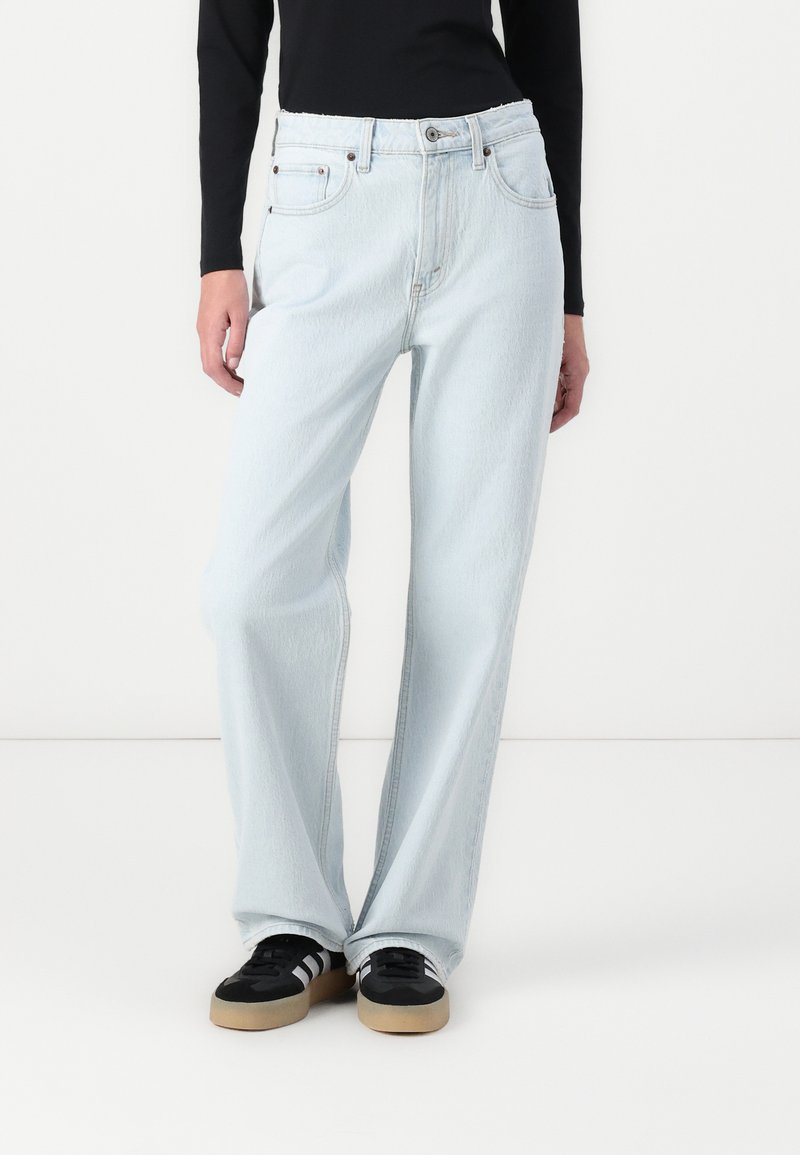 Jean clair taille haute avec une coupe loose et large, avec des poches avant et des passants de ceinture, associés à des chaussures noires.