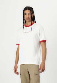 White cotton t-shirt with red crew neck and sleeve cuffs, featuring a cursive red logo on the front. Model wears beige cargo shorts.