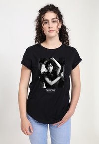 Woman with curly hair wearing black "Wednesday" T-shirt featuring a black-and-white image of a girl dancing with raised arms.