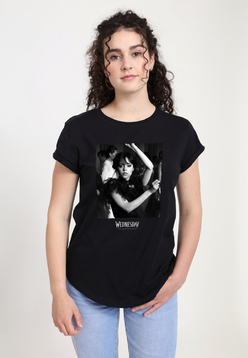 Woman with curly hair wearing black "Wednesday" T-shirt featuring a black-and-white image of a girl dancing with raised arms.