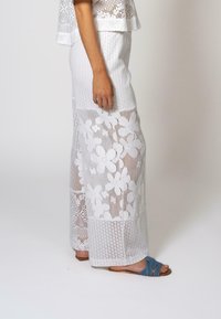 Pantalons large en dentelle blanche avec des motifs floraux et un design texturé, associés à des sandales bleues à brides croisées.