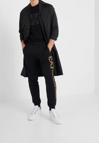 Svart trenchcoat över en svart t-shirt med logotyp, ihop med svarta sweatpants med guldtext och vita sneakers.